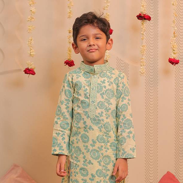 A young boy wearing a light green and cream printed kurta with beige trousers, standing in front of a decorated backdrop with hanging garlands and cushions.