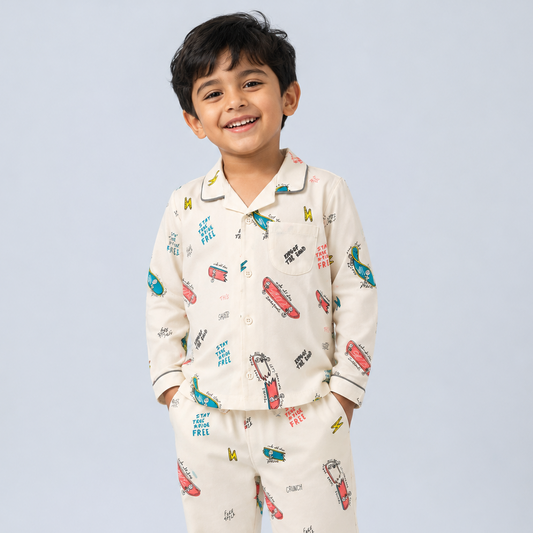 A young boy smiling and wearing a two-piece pajama set with a cream base and a colorful cartoon print of cars, buses, and airplanes, paired with white sneakers, set against a light gray studio background.