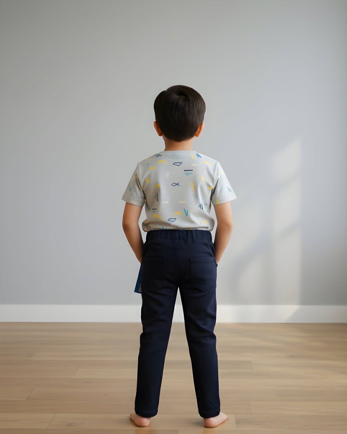 Child wearing a patterned shirt and dark pants standing in a room with a plain wall and wooden floor.