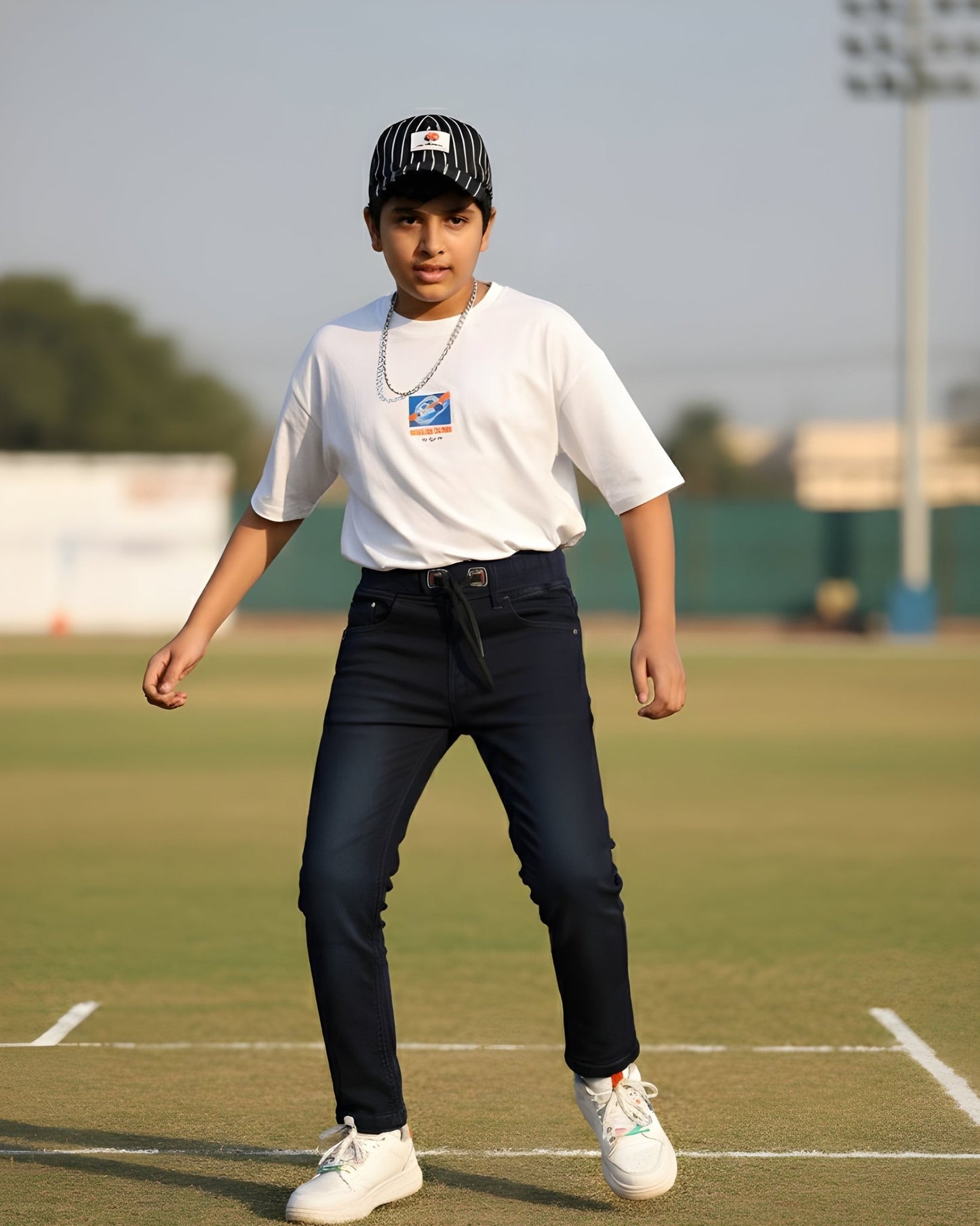 Person standing on a sports field wearing a white t-shirt, dark pants, and a cap.
