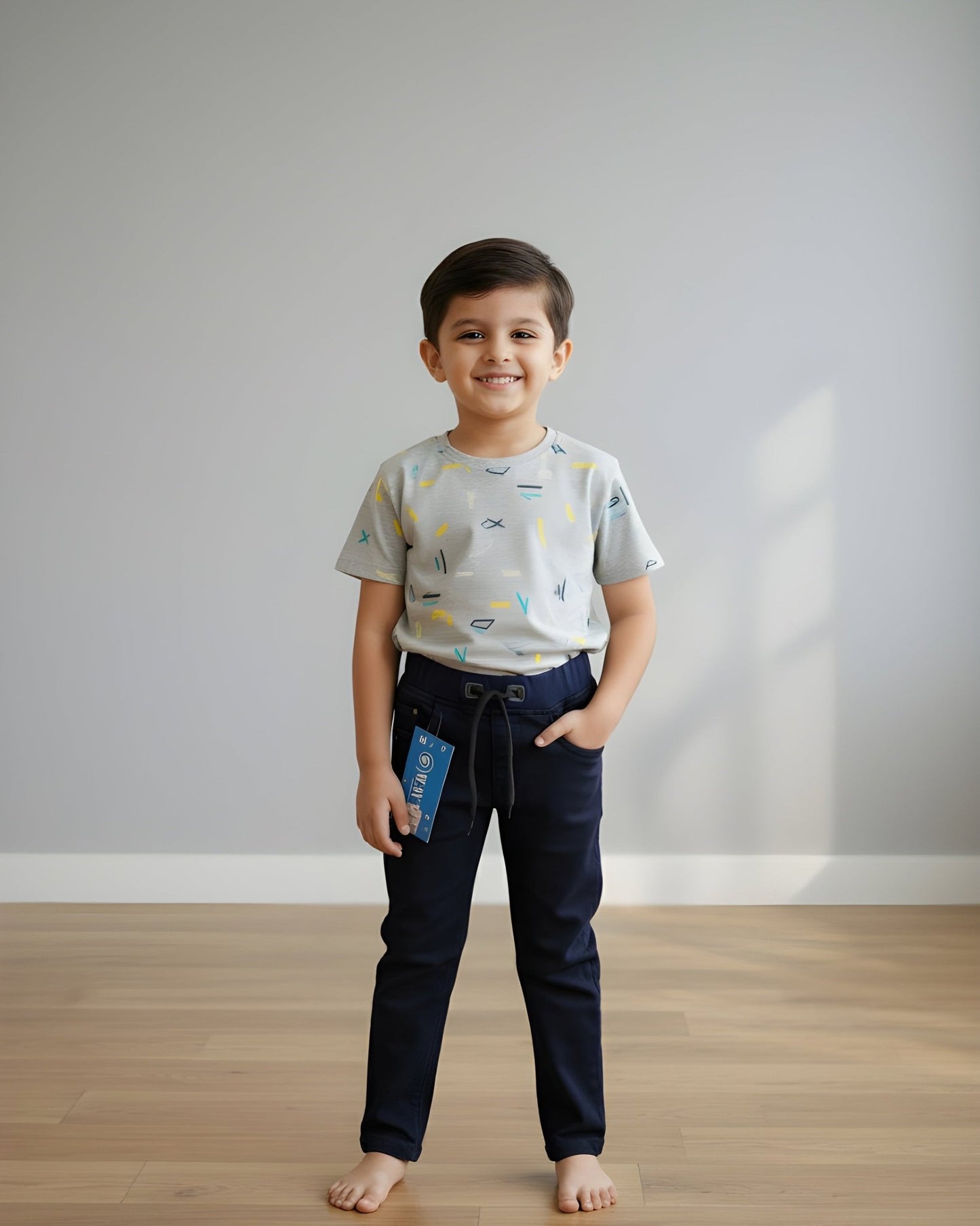 Child wearing a patterned shirt and dark pants standing on a wooden floor with a plain white wall background