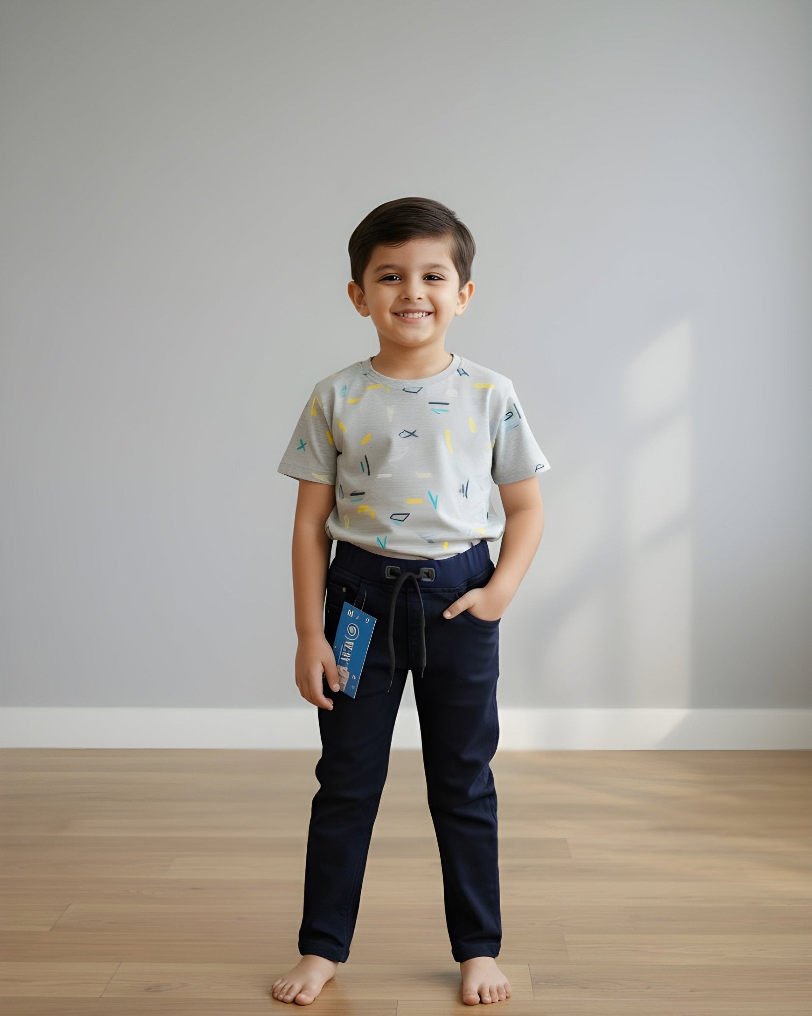 Child wearing a patterned shirt and dark pants standing on a wooden floor with a plain white wall background